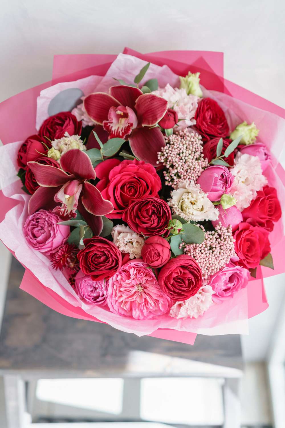 Bright red bouquet of beautiful flowers on wooden table. Floristry concept. Spring colors. the work of the florist at a flower shop. Vertical photo Crimson Love Bouquet with red and hot pink roses, cymbidium orchids, lisianthus, pink berries, and greenery wrapped in vibrant pink paper
